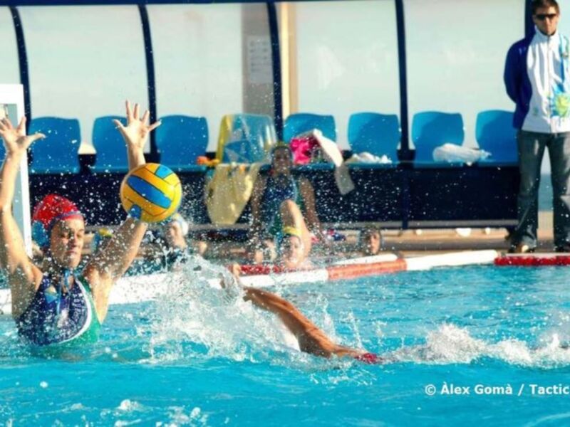 Jugadores de waterpolo en acción durante un partido, con un balón en el aire en un estadio cubierto.