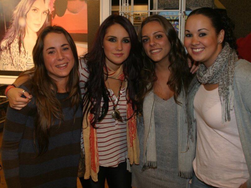 Las cuatro chicas son sonrientes y están posando para una foto. La imagen en el fondo parece ser de una celebridad.