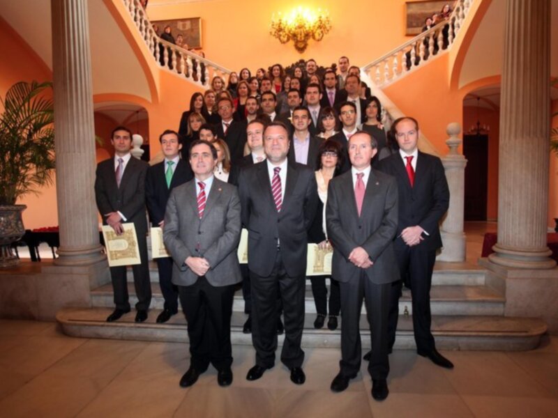 Grupo de personas en trajes formales posando en un interior clásico con escalera y columnas.