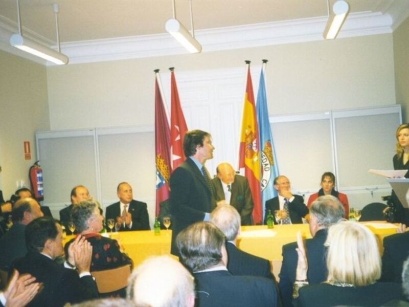 Una reunión formal en una sala con mesas y personas sentadas, algunas personas están levantando sus manos para saludar.