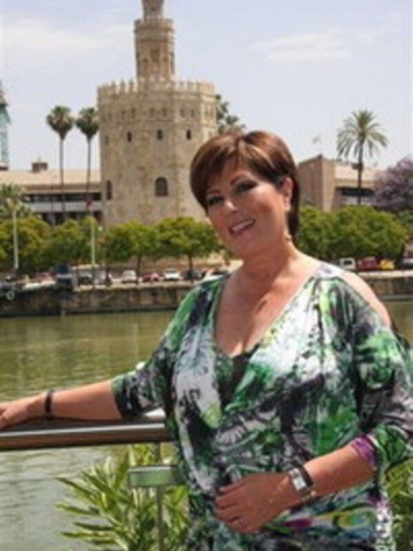 A una persona frente al famoso Torre del Oro de Sevilla, España. La torre se destaca en el fondo, con su estructura clásica y color dorado. La persona está posando frente al río, con un vestido verde que combina bien con el entorno.