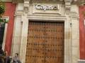 Una entrada principal de un edificio con el nombre "Cajasol" en letras grandes sobre la puerta. La fachada es de piedra y tiene un balcón con banderas rojas en el primer piso. La puerta principal es de madera y tiene detalles metálicos.