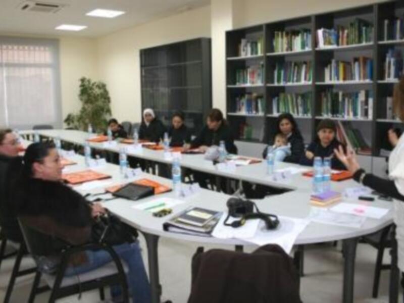 Una clase de trabajo en una sala de conferencias con estudiantes sentados en mesas y una profesora en la parte frontal explicando algo.