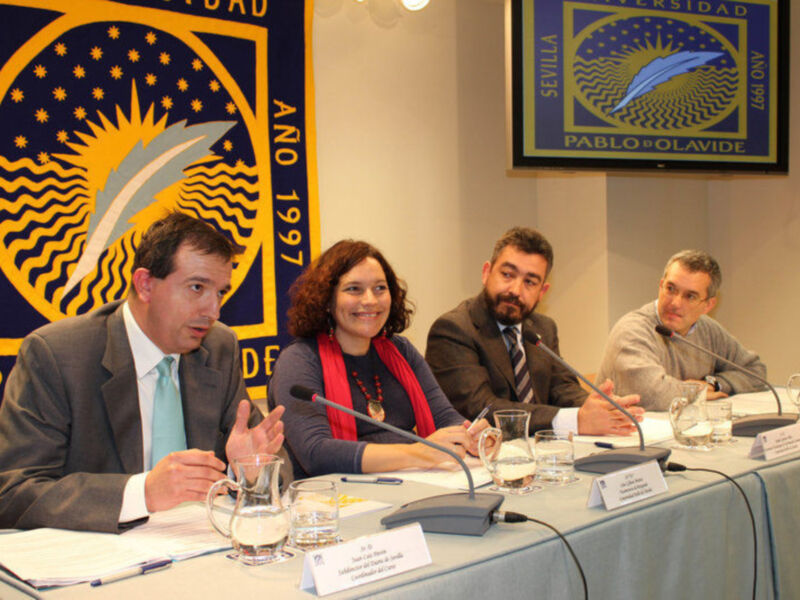 Cuatro personas sentadas en una mesa, dos hombres y dos mujeres, con micros en frente de ellos. Detrás hay una bandera con el nombre 'Pablo Olavide' y otro cartel con un símbolo de sol y estrellas.