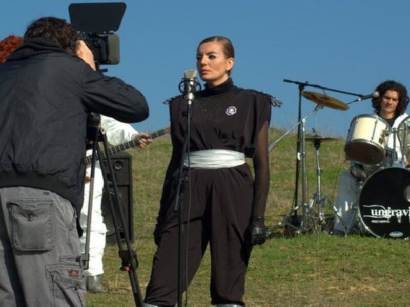 Una mujer con una chaqueta negra y pantalones negros está en el centro de la imagen, con un micrófono en frente de ella. A su derecha, hay una batería y a su izquierda, un hombre con una cámara de video está filmando. En el fondo, se puede ver a otra persona tocando la guitarra y un hombre con una camisa azul está tomando fotos. La escena parece ser en un campo o jardín con césped y cielo azul.