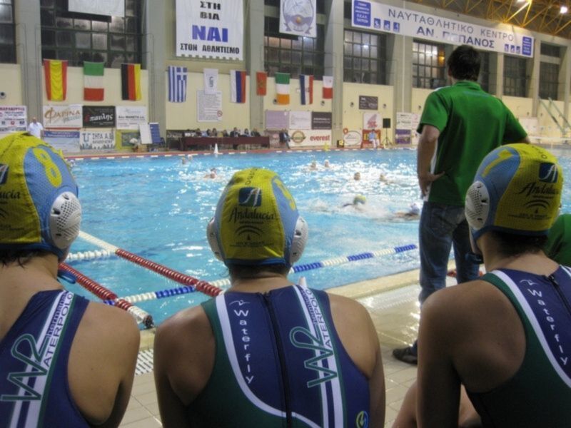 Competencia de waterpolo en una piscina cubierta, con nadadores en trajes y cascos de equipo.
