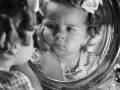 Una niña mira su reflejo en un espejo redondo, con una peluca y una corbata de lazo en el cabello. La imagen es blanco y negro, capturando un momento de introspección infantil.