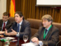 Tres hombres en traje se encuentran sentados en una mesa de conferencia, con un fondo que incluye una bandera española y una pantalla.