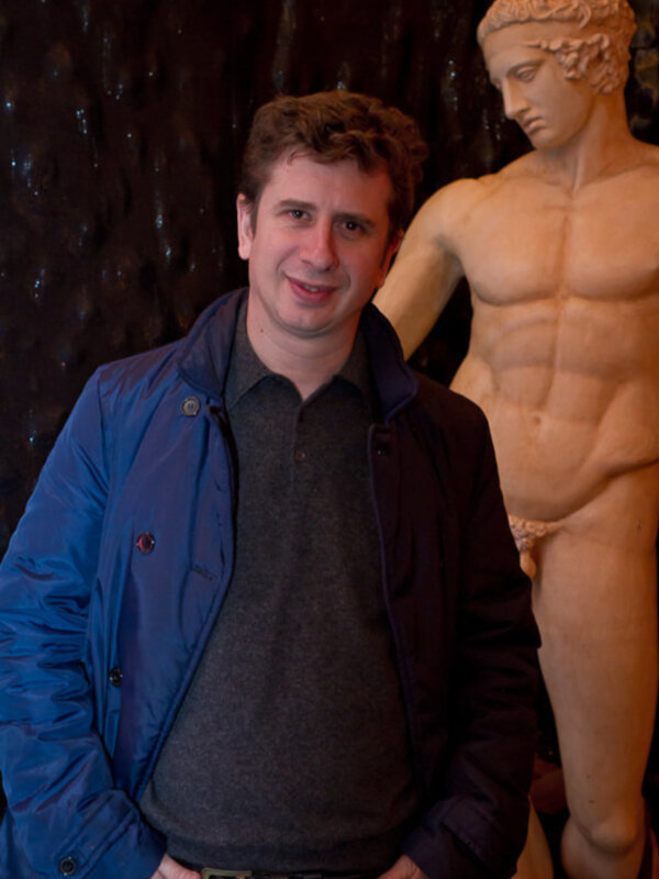 Un hombre en una chaqueta azul posa junto a una estatua de un atleta musculoso, posiblemente en un museo.