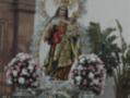 Una estatua de la Virgen María con un niño en sus brazos, rodeada por flores y decoraciones. La imagen se encuentra frente a una estructura arquitectónica con plantas en primer plano.