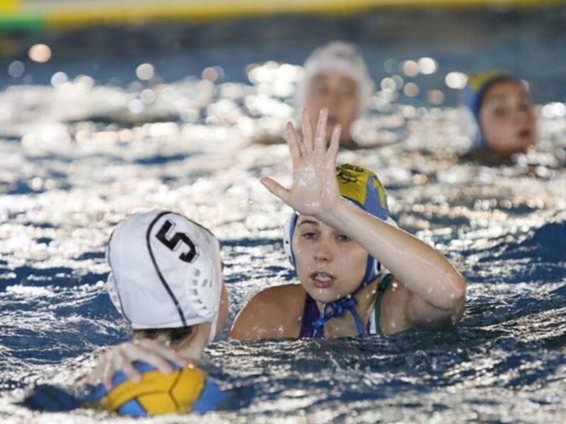 Jugadoras de waterpolo en acción durante un partido, con una jugadora levantando la mano en el agua.