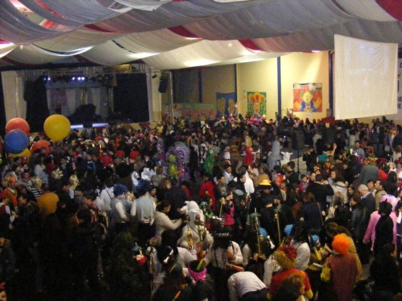 Fiesta vibrante con multitud de personas disfrutando en un evento festivo bajo una estructura decorativa con globos y telón de fondo.