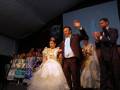 Una boda tradicional en un escenario con una novia vestida de blanco, un padrino y otros invitados en trajes formales.