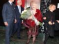 Una mujer vestida con un abrigo rojo y falda de lentejuelas camina con una gran florero en la mano, seguida por tres hombres vestidos formalmente. La escena parece ser una celebración o evento importante en un entorno urbano con luces rojas y un edificio de fondo.