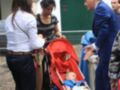 Un hombre en traje azul habla con una mujer y un niño en un carrito rojo. Detrás de ellos, hay otros dos adultos conversando y una persona con un vestido azul. La escena parece ser en un lugar público, posiblemente una calle o plaza.