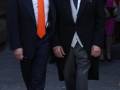 Dos hombres vestidos formalmente caminan por una calle, uno con corbata naranja y el otro con un elegante traje negro. La imagen sugiere una ceremonia o evento importante.