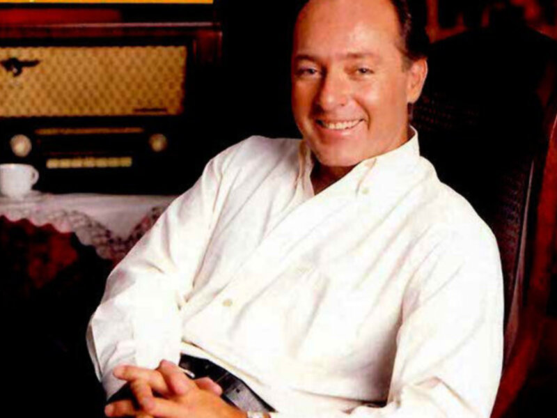 Un hombre sonríe ampliamente mientras se sienta en una silla. Está vestido con un elegante abrigo blanco y tiene los brazos cruzados sobre el pecho. En la mesa a su lado, hay una taza de café y un estéreo antiguo. La escena transmite un ambiente relajado y cómodo, probablemente en una sala de estar.