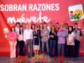 Una foto de un grupo de personas en una presentación, con un fondo rojo que dice "Sobran razones muévete" y una imagen de un sol en el logo. La gente está posando frente a flores rojas y una bandera.