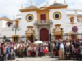 Fotografía de una multitud reunida frente a la plaza de toros de Sevilla, España. La imagen muestra un edificio histórico con detalles arquitectónicos españoles y una multitud de personas, algunas sosteniendo paraguas. La escena parece ser parte de una celebración o evento importante en la ciudad.