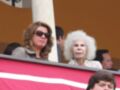 Dos mujeres, una con gafas de sol y otra con cabello blanco, observan desde un balcón. El fondo es una pared de color naranja con una bandera roja y blanca en primer plano.