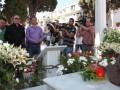 Una multitud se reúne en un cementerio para rendir homenaje a una persona fallecida. Los asistentes, algunos con cámaras de fotos, se concentran en el ataúd cubierto de flores blancas y rojas. La escena conmemora un evento trágico, capturando el dolor y la resiliencia de los presentes.