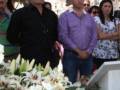 Una imagen de una ceremonia funeraria con flores blancas y rosadas en el centro, rodeada por personas en traje de funeral.