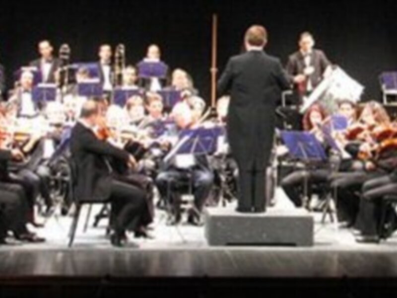 Orquesta sinfónica en concierto en un escenario de teatro, con músicos tocando instrumentos de cuerda y viento.