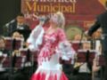 Una mujer cantante en traje de flamenca roja y blanco, frente a un grupo de músicos bajo una pancarta que dice "Banda Sinfónica Municipal de Sevilla".