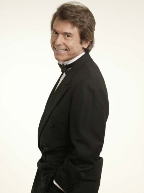 Portrait of a smiling man in a black tuxedo, standing against a white background.