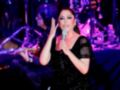 Una cantante en escenario con un vestido negro brillante, sosteniendo un micrófono y gesturando con las manos.
