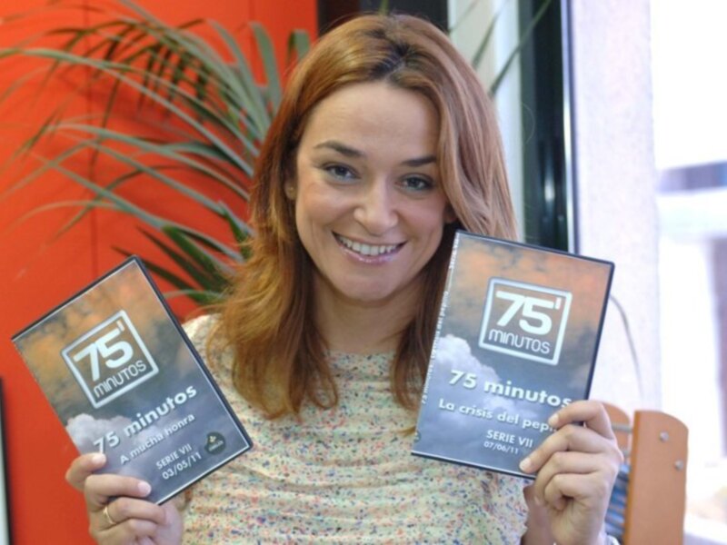 Una mujer sonríe mientras sostiene dos DVDs de la serie '75 minutos' en un entorno acogedor con plantas y paredes de color rojo.