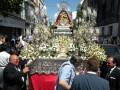 Imagen de una procesión religiosa con una imagen venerada en un carro decorado con flores y luces, rodeada por fieles observando la celebración.