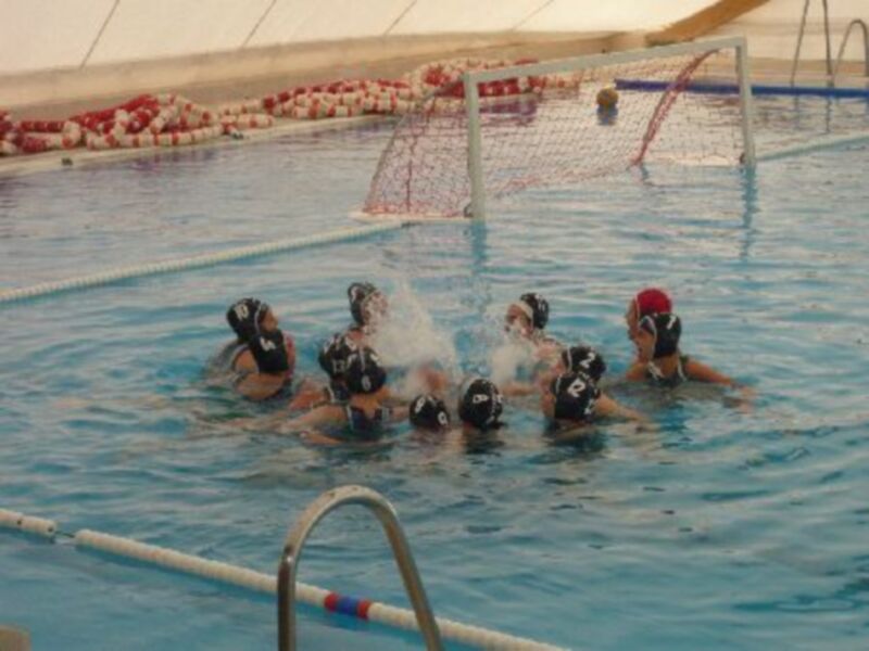 Un grupo de jugadoras de waterpolo en acción en una piscina. La imagen muestra a las jugadoras con cascos y uniformes, jugando en un campo de waterpolo. La acción está centrada alrededor del portero, que está en el área de gol. El agua es clara y la piscina tiene una red de portería visible en el fondo.