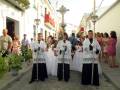 Un grupo de personas participa en una procesión religiosa. Varias personas vestidas con trajes blancos y negros, posiblemente de una tradición o festividad local, caminan por una calle con edificios blancos y rojos. La imagen muestra un ambiente festivo, posiblemente durante una celebración religiosa o cultural.