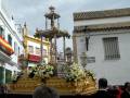 Un carro procesional dorado con flores blancas y amarillas, flanqueado por edificios de colores en una calle típica andaluza.