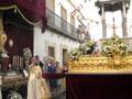 Procesión religiosa con sacerdote oficiando frente a un paso ornado con flores y mantas rojas.