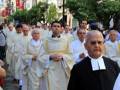 Un grupo de sacerdotes vestidos con hábitos blancos y negros caminan por una calle con edificios históricos al fondo. Banderas ondean en el aire, y algunos de los sacerdotes llevan gafas.