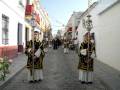 Procesión religiosa en una calle adoquinada, con personas vestidas con trajes tradicionales y portando estandartes.