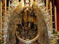 Imagen religiosa de una Virgen María vestida con ornamentos dorados y plateados, rodeada de velas y flores, en un entorno de altar.