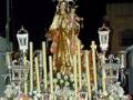 Estatua de la Virgen con el Niño en una procesión nocturna, rodeada de flores y luces.