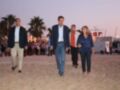 Un grupo de personas caminan por la playa, con algunos hombres vestidos formalmente y una mujer en jeans. En el fondo, se pueden ver palmeras y un edificio con letras grandes.