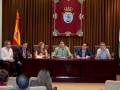 Una reunión formal en un salón con una bandera nacional, un escudo heráldico y varios participantes sentados en sillas.