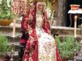 Una figura vestida con una túnica roja y blanca, posiblemente en un entorno religioso o ceremonial. La túnica está adornada con detalles dorados y el personaje lleva una corona en la cabeza. La escena parece ser dentro de un templo o iglesia, con elementos decorativos y una mesa en el fondo.