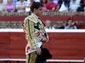 Un torero en traje tradicional de color dorado y verde, con un sombrero negro, camina por el ruedo en una plaza de toros. La audiencia se puede ver en los asientos del fondo, con algunos espectadores tomando fotos.