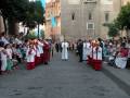 Un grupo de personas vestidas con trajes tradicionales se reúnen en una calle adoquinada, frente a un edificio histórico con torres. La gente observa atentamente mientras los individuos en trajes se preparan para una ceremonia o celebración.