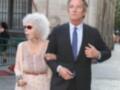 Una anciana con cabello blanco y gafas de sol, camina junto a un hombre en traje formal. Ambos parecen estar en una calle con edificios de fondo.