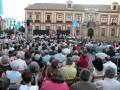 Una multitud asiste a un concierto en una plaza pública frente a un edificio histórico con arcos y ventanas. La escena está iluminada por la tarde, con un cielo azul y nubes dispersas. La gente está sentada en gradas, disfrutando del evento musical.