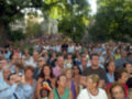 Una multitud en un parque, con personas tomando fotos y disfrutando del ambiente. En el fondo, se ve un monumento con una estatua y banderas ondeando.
