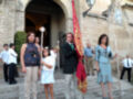 Una familia participa en una procesión religiosa. La madre sostiene un bastón, la hija un palo y el padre una bandera roja con detalles dorados. Todos están vestidos formalmente, frente a un edificio histórico con una puerta abierta.