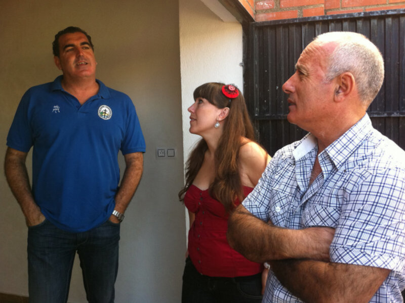 Un hombre en una camiseta azul y otro en una camisa a cuadros charlan mientras una mujer en un vestido rojo con un accesorio en el cabello observa la conversación.